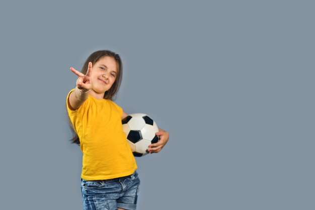 Une joueuse de sport FFan tient un ballon de football en fête, un enfant souriant heureux en t-shirt jaune sur fond gris, montre le geste de la victoire, espace de copie de texte gratuit