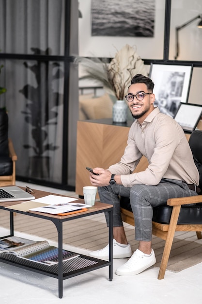 Jour de chance. Homme séduisant et réussi dans des verres avec un smartphone souriant à la caméra assis dans un fauteuil près de la table avec un ordinateur portable