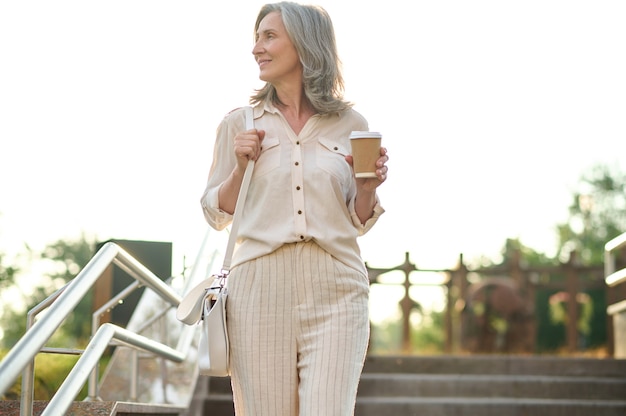 Jour de chance. Jolie femme élégante adulte avec du café à côté dans les escaliers dans le parc le jour d'été