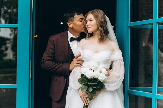 Jour du mariage Belle mariée européenne et son marié asiatique posant sur fond de cafés et de grandes fenêtres