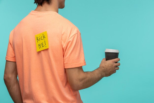 Jour du poisson d'avril mec par derrière autocollant post-it jaune kick me tshirt orange tenant une tasse de café