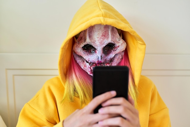 Jour d'Halloween, une fille au masque effrayant de cauchemar avec une capuche sur la tête fait un appel vidéo sur un smartphone, s'assoit sur le lit à la maison, fête d'halloween