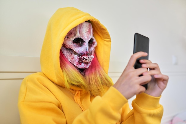 Jour d'Halloween, une fille au masque effrayant de cauchemar avec une capuche sur la tête fait un appel vidéo sur un smartphone, s'assoit sur le lit à la maison, fête d'halloween