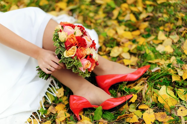 Jour de mariage. Gros plan des pieds de la mariée en chaussures rouges, bouquet de mariée coloré et feuilles d'automne sur l'herbe verte