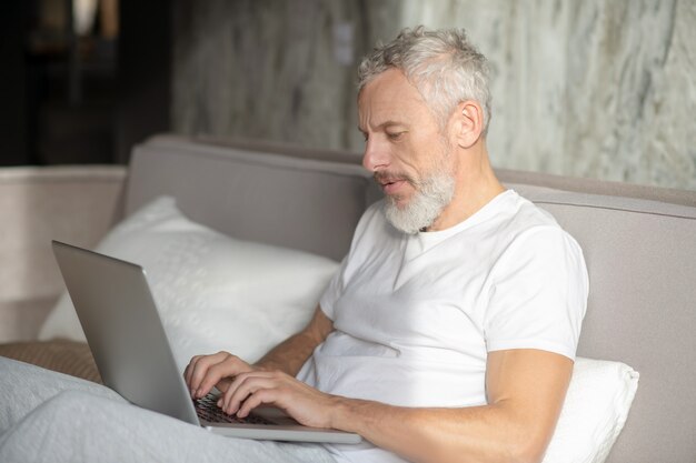 Jour de repos. Homme aux cheveux gris sérieux de profil travaillant sur ordinateur portable s'appuyant sur des oreillers dans son lit à la maison