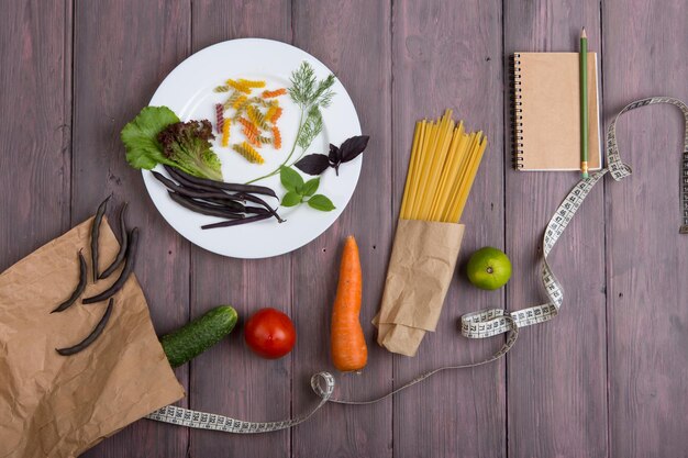 Journal alimentaire diététique - cahier pour compter les calories, ruban centimétrique, légumes frais dans un sac en papier écologique et sur assiette, pâtes spaghetti sur table en bois