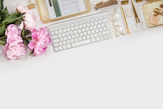 Journal des femmes et papeterie dorée. Bouquet de pivoines roses. Des lunettes, un clavier blanc, un stylo, des ciseaux et du café sur le bureau.