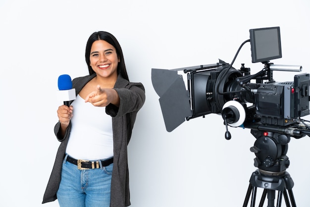 Journaliste colombienne femme tenant un microphone et rapportant des nouvelles sur le mur blanc faisant un geste de téléphone et pointant vers l'avant