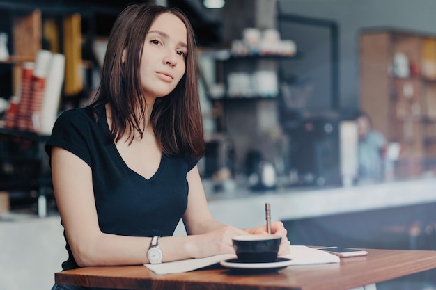 Une journaliste concentrée pense à un nouvel article écrit des informations dans le bloc-notes boit une boisson aromatique chaude dans un café un téléphone intelligent moderne sur une table près du processus de travail