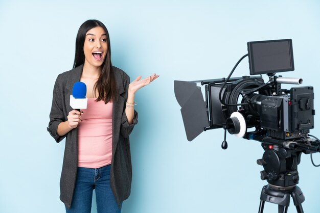 Journaliste femme tenant un microphone et rapportant des nouvelles sur le mur bleu avec une expression faciale surprise