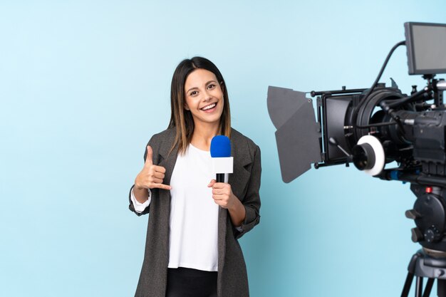 Journaliste femme tenant un microphone et rapportant des nouvelles sur le mur bleu faisant un geste de téléphone