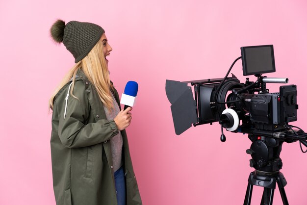 Journaliste femme tenant un microphone et rapportant des nouvelles sur le mur rose en riant en position latérale