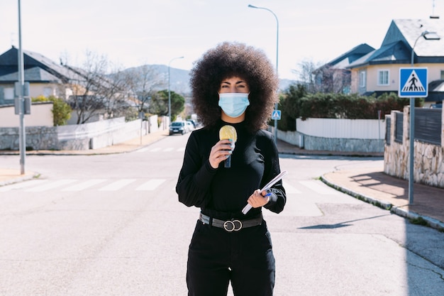Journaliste de télévision aux cheveux afro, portant un masque chirurgical pour se protéger d'un virus, rapportant une histoire dans la rue