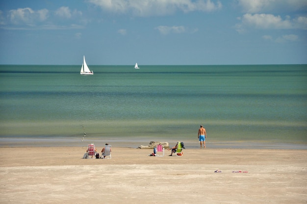 Journée ensoleillée sur une plage avec une mer calme et des voiliers à l'horizon Certaines personnes prennent le soleil sur le sable