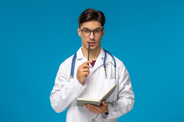 Journée des médecins beau mec mignon brune en robe médicale mordre un stylo et tenant un livre