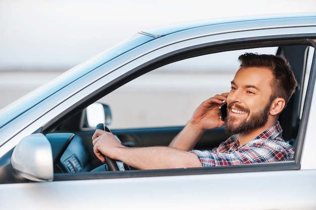 La journée parfaite pour conduire. Vue latérale d'un jeune homme gai conduisant sa voiture et parlant au téléphone portable