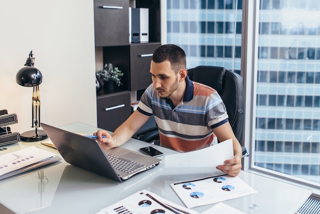 Journée de travail d'analyste financier tapant sur ordinateur analysant le travail avec des statistiques assis sur le lieu de travail contre la fenêtre avec vue sur le paysage urbain.