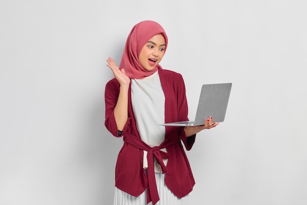 Joyeuse belle femme asiatique en chemise décontractée et hijab utilisant un ordinateur portable, faisant un appel vidéo, faisant des gestes de salut à des amis isolés sur fond blanc. Concept de mode de vie religieux de personnes