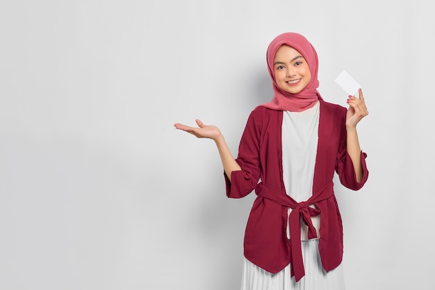 Joyeuse belle femme asiatique dans une chemise décontractée et hijab montrant une carte de crédit, présente un espace vide à la main isolé sur fond blanc. Concept de mode de vie religieux de personnes