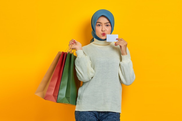 Joyeuse belle femme asiatique en pull blanc tenant des sacs à provisions et une carte de crédit isolée sur fond jaune