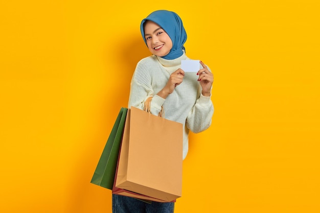 Joyeuse belle femme asiatique en pull blanc tenant des sacs à provisions et montrant une carte de crédit isolée sur fond jaune