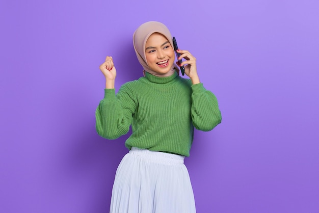 Joyeuse belle femme asiatique en pull vert parlant de téléphone portable tout en se réjouissant de la chance isolée sur fond violet