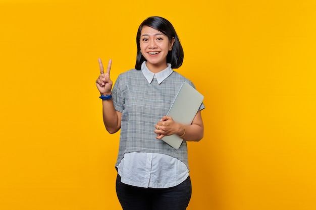Joyeuse belle femme asiatique tenant un ordinateur portable et montrant un signe de paix sur fond jaune