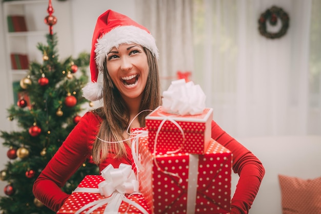 Joyeuse belle jeune femme portant un bonnet de Noel et tenant de nombreux cadeaux de Noël à la maison.