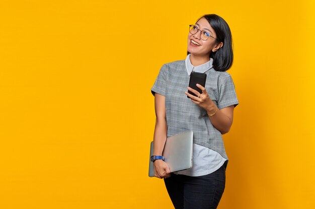 Joyeuse belle jeune femme regardant l'écran du téléphone portable et tenant un ordinateur portable sur fond jaune