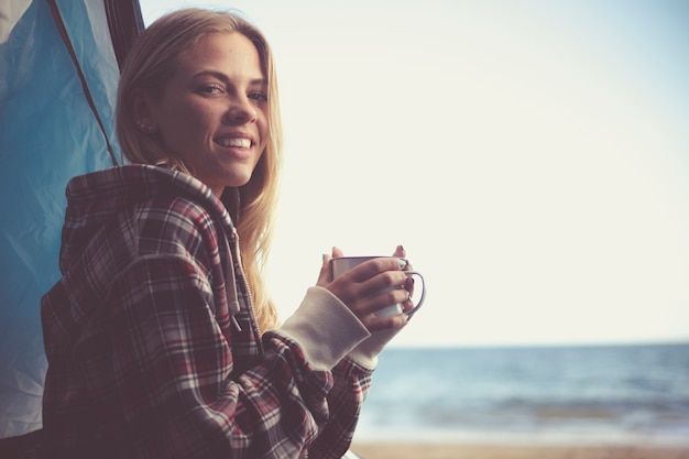 Joyeuse belle jeune fille blonde voyageuse tient une tasse de café ou de thé à l'intérieur de sa tente campant à la plage avec liberté et vue sur l'océan