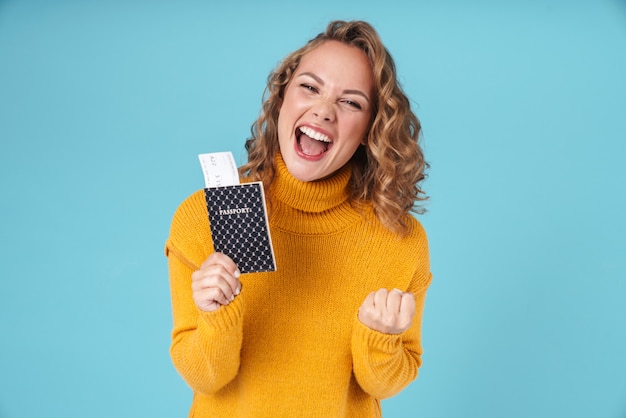 Joyeuse belle jeune fille portant un chandail debout isolé sur bleu, montrant un passeport et des billets d'avion