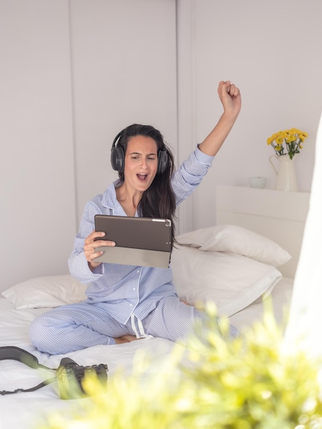 Joyeuse brune en vêtements de nuit assise sur le lit et portant des écouteurs tout en levant le bras et en regardant la tablette à la main