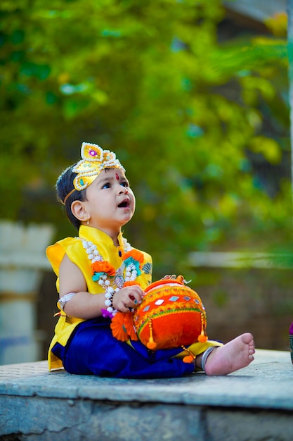 Joyeuse carte de voeux Janmashtami montrant un petit garçon indien se faisant passer pour Shri krishna ou kanha/kanhaiya avec une photo de Dahi Handi et des fleurs colorées.