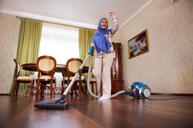 Joyeuse charmante jeune femme arabe musulmane, femme au foyer, femme de ménage avec la tête couverte de hijab bleu aspirant le sol, dansant et chantant pendant le nettoyage de la maison