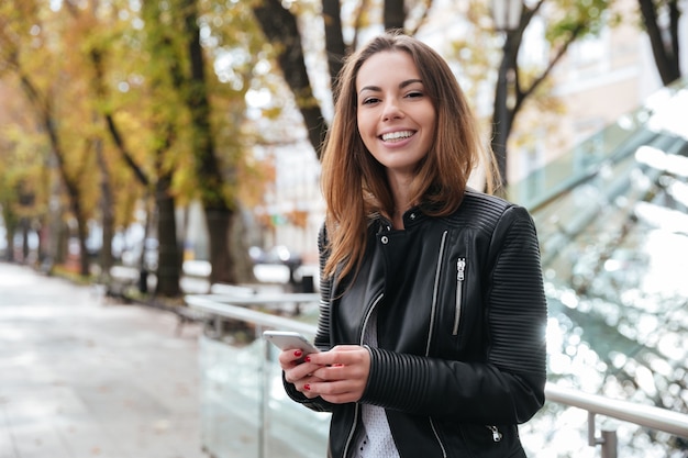 Joyeuse charmante jeune femme debout et utilisant un smartphone dans la ville