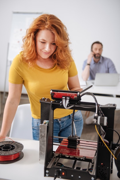 Joyeuse créatrice professionnelle debout devant une imprimante 3D et vérifiant ses capacités techniques tout en tenant une tablette