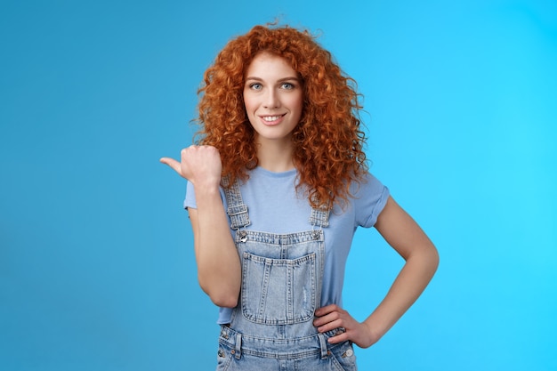 Joyeuse curieuse rousse animée belle fille aux cheveux bouclés portant un t-shirt d'été pointant le pouce gauche regarder la caméra intriguée excitée explorer de nouveaux magasins amis marchant dans un centre commercial.