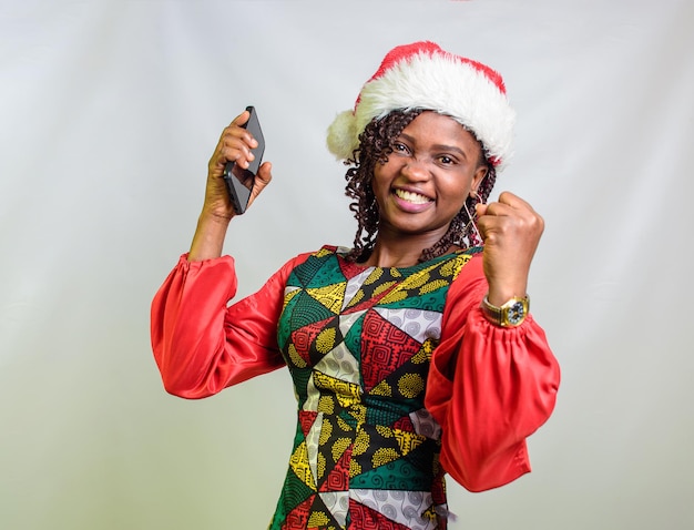 Une joyeuse dame africaine du Nigeria tenant un téléphone intelligent et ayant également une casquette de Noël sur la tête