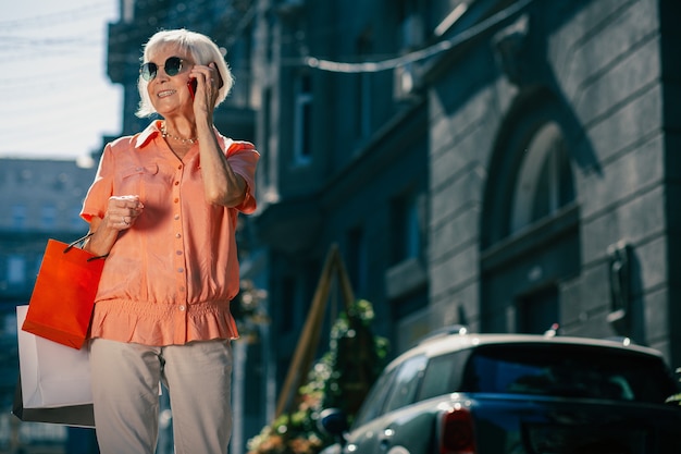 Joyeuse dame âgée avec des sacs en papier colorés dans la rue ayant une conversation téléphonique agréable et souriant