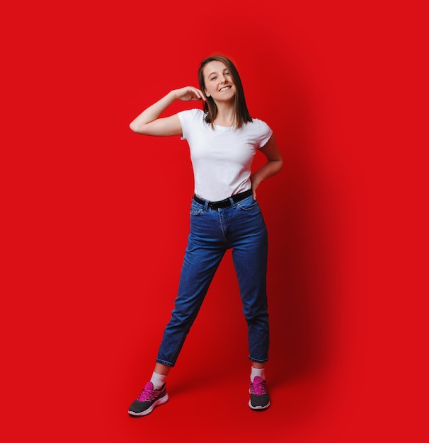 Joyeuse dame de race blanche vêtue d'un t-shirt blanc et d'un jean bleu touche ses cheveux et pose sur fond rouge avec un espace vide