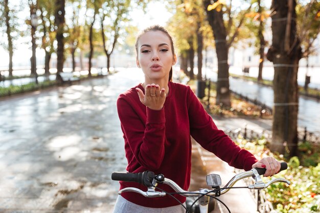 Joyeuse dame vêtue d'un pull marchant avec son vélo à l'extérieur tout en faisant un geste OK