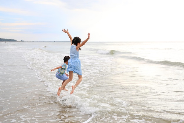 Photo joyeuse enfant asiatique fille et petit garçon s'amusant à sauter sur la vague à la plage de sable tropicale au coucher du soleil bonne famille profiter des vacances d'été