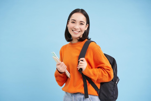 Joyeuse étudiante asiatique avec sac à dos et cahiers allant au collège semble heureux fond bleu