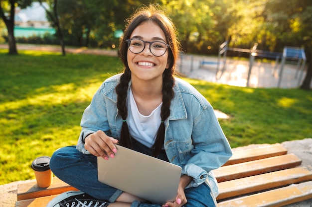 Joyeuse étudiante assise sur un banc dans le parc, étudiant sur un ordinateur portable