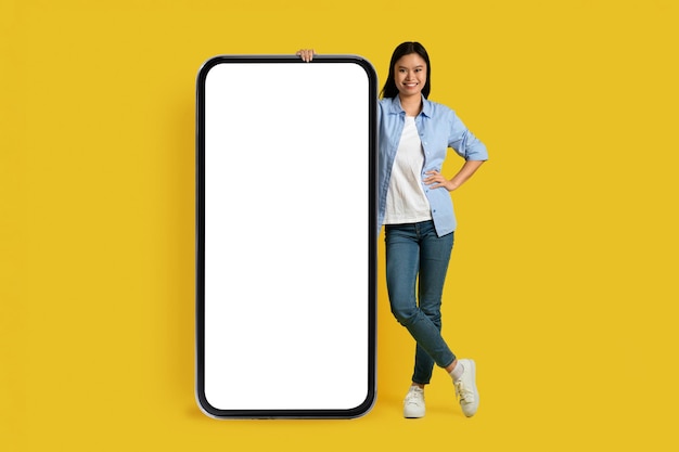 Joyeuse étudiante coréenne du millénaire dans un stand décontracté avec un énorme smartphone avec écran vide