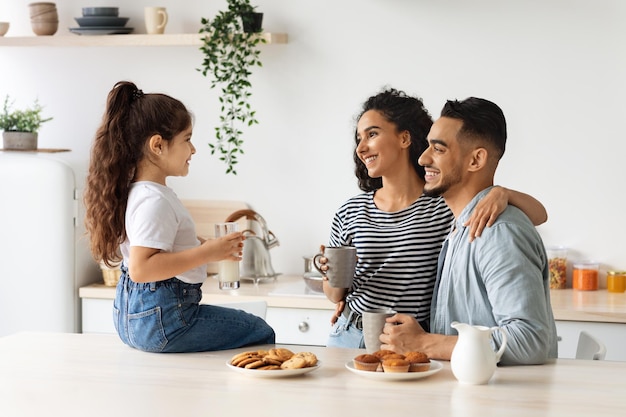 Joyeuse famille arabe prenant son petit-déjeuner dans une cuisine confortable, petite fille assise sur une table, buvant du lait et discutant avec un père et une mère souriants qui s'embrassent en buvant du café du matin