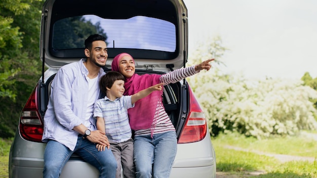 Joyeuse famille arabe de trois personnes assises dans un coffre de voiture ouvert et pointant de côté, parents musulmans heureux et enfant mâle mignon passant du temps à l'extérieur, profitant de voyages d'été, panorama avec espace de copie