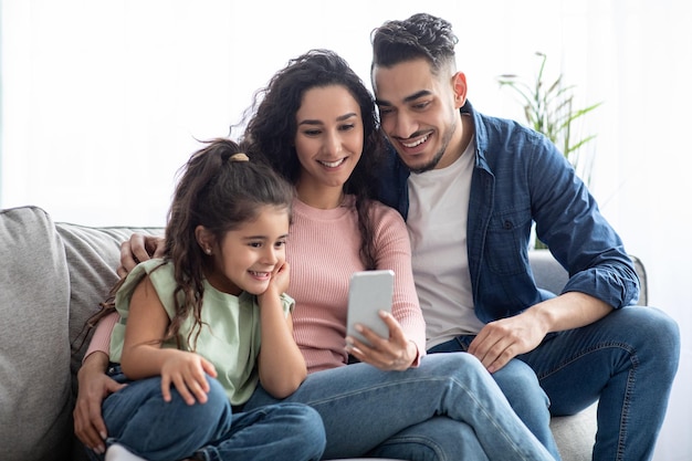 Joyeuse famille arabe de trois personnes utilisant un smartphone à la maison, de jeunes parents et leur jolie petite fille regardant des vidéos ou naviguant sur Internet tout en se relaxant sur un canapé dans le salon ensemble, en gros plan