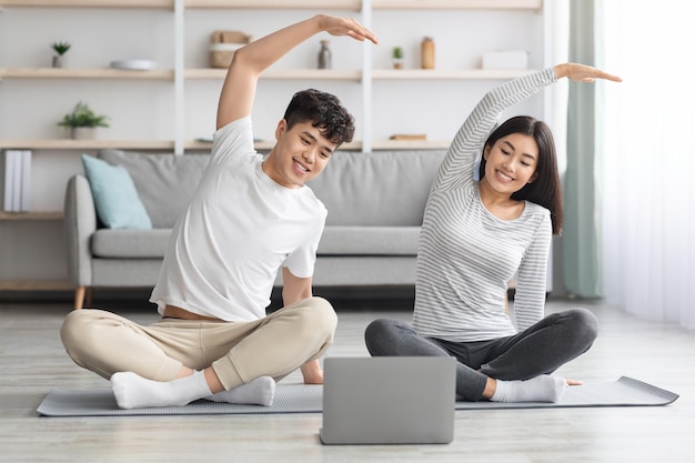 Joyeuse famille asiatique pratiquant le yoga à la maison, s'étirant sur un tapis de fitness, regardant un écran d'ordinateur portable, ayant des cours en ligne, un intérieur de salon, un espace de copie. Couple noir sportif exerçant ensemble