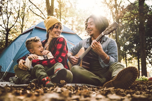 Joyeuse famille campant dans la forêt jouant de la guitare et chantant ensemble - Mère, père et fils s'amusant à faire du trekking dans la nature assis devant la tente - Concept famille, nature et trekking